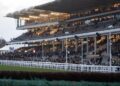 Packed grandstands at Cheltenham Racecourse at dusk, seen from across the track with a fence in the foreground