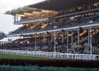 Packed grandstands at Cheltenham Racecourse at dusk, seen from across the track with a fence in the foreground